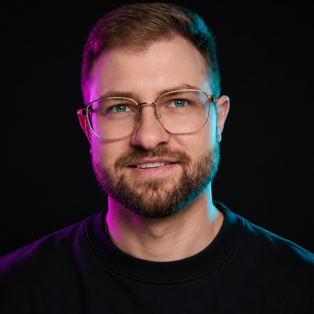 Studio portrait of a smiling person with glasses, short beard, and black shirt on a dark background with pink and blue lighting