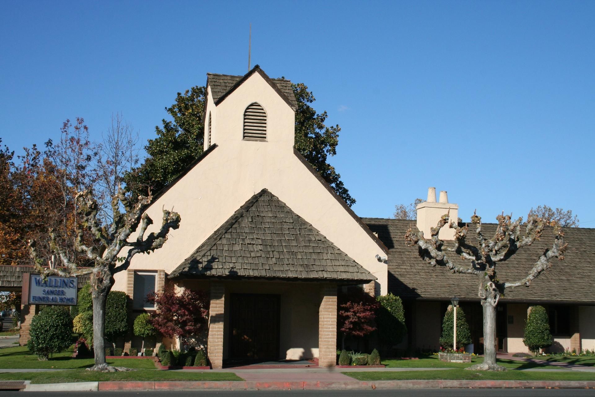 Small white chapel with a steep roof, framed by trees and shrubs under a clear blue sky.
