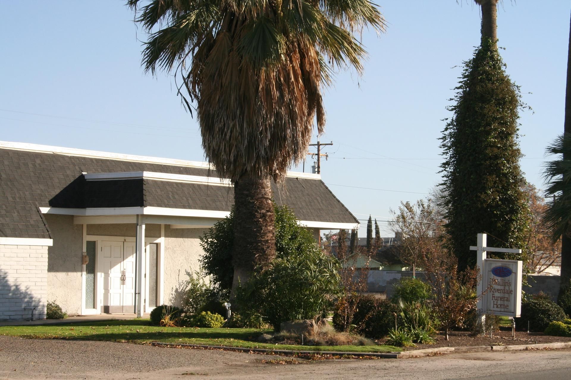 White building with black awning, tall palm trees, and a roadside sign under a clear sky