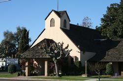Small beige church with dark shingled roof and a bell tower, surrounded by trees under a blue sky