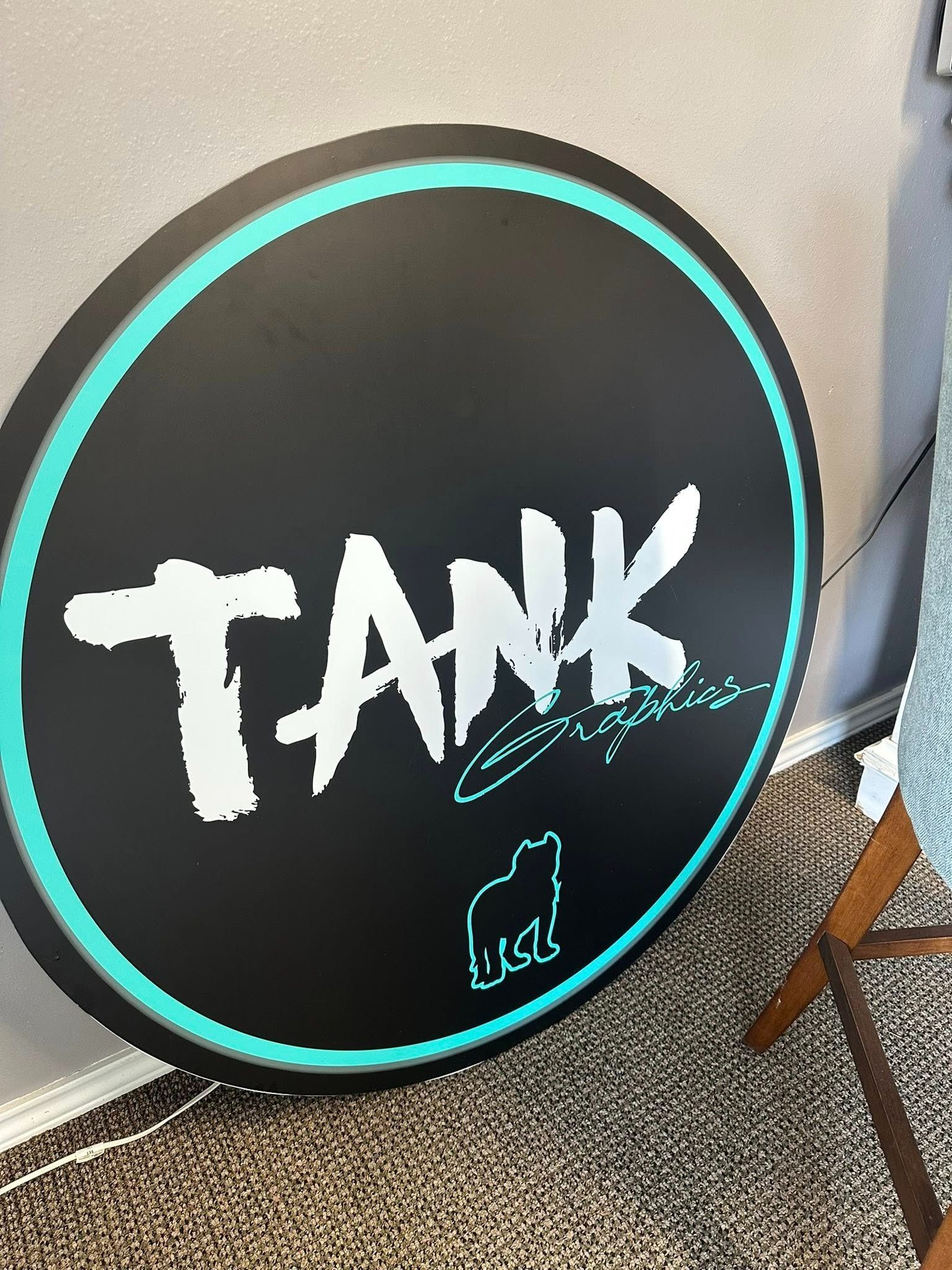 Round black sign with teal rim and “TANK” in white graffiti lettering on a carpeted floor.
