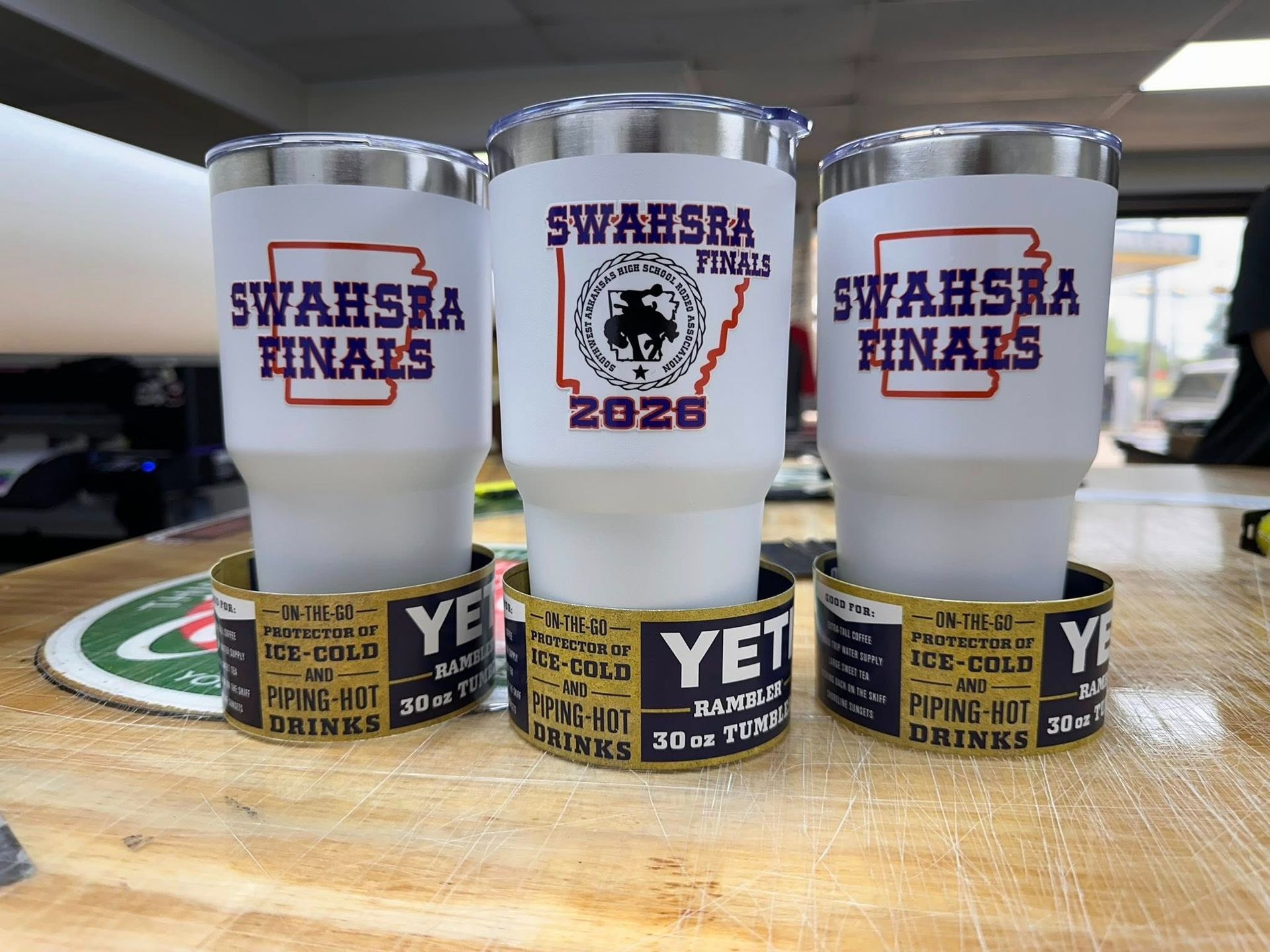 Three white Yeti tumblers with purple and gold event logos on a wooden table in an office.