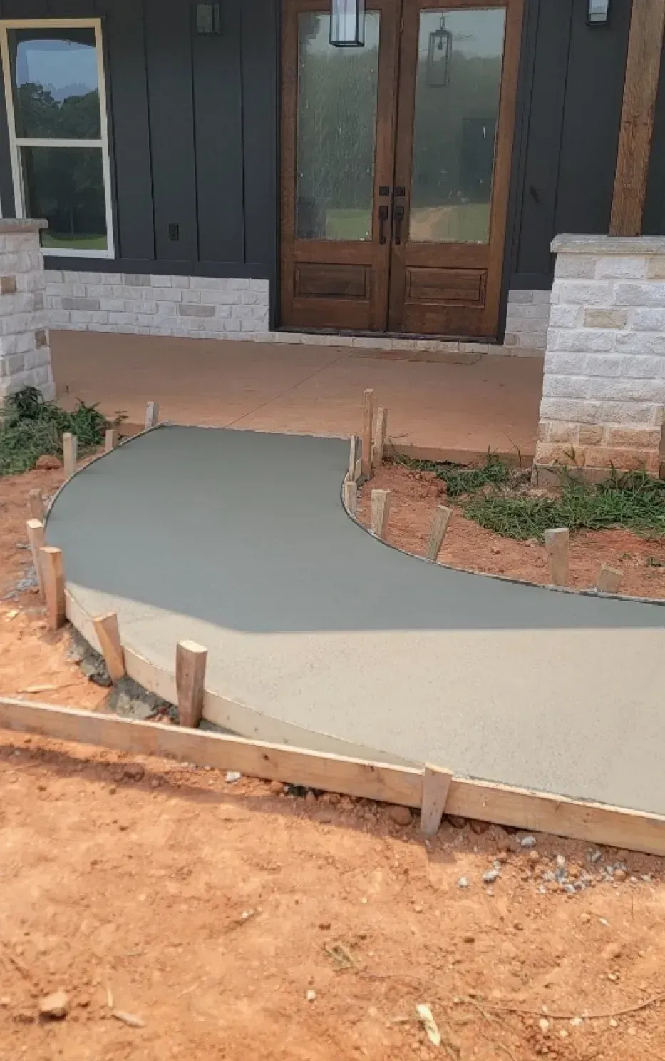 A fresh, curved concrete walkway leads to the double wooden front doors of a house with stone-accented exterior walls.