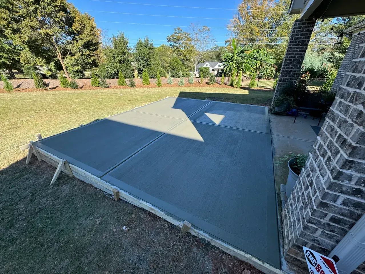 A freshly poured concrete patio slab with wooden formwork extends from a brick house into a grass yard on a sunny day.