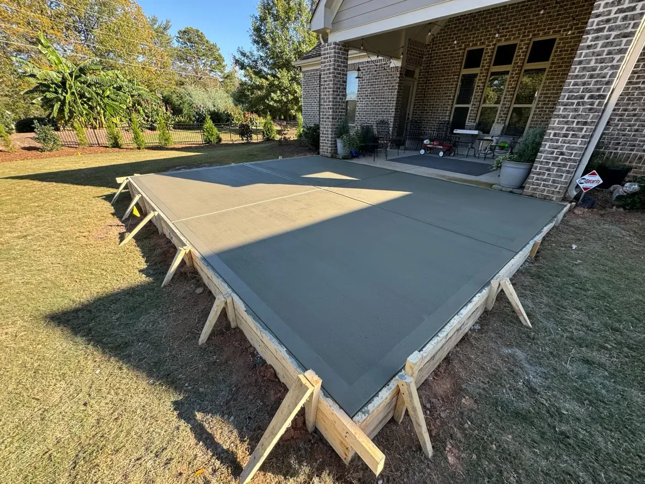A newly poured rectangular concrete patio slab held in place by wooden forms, situated next to a brick house.