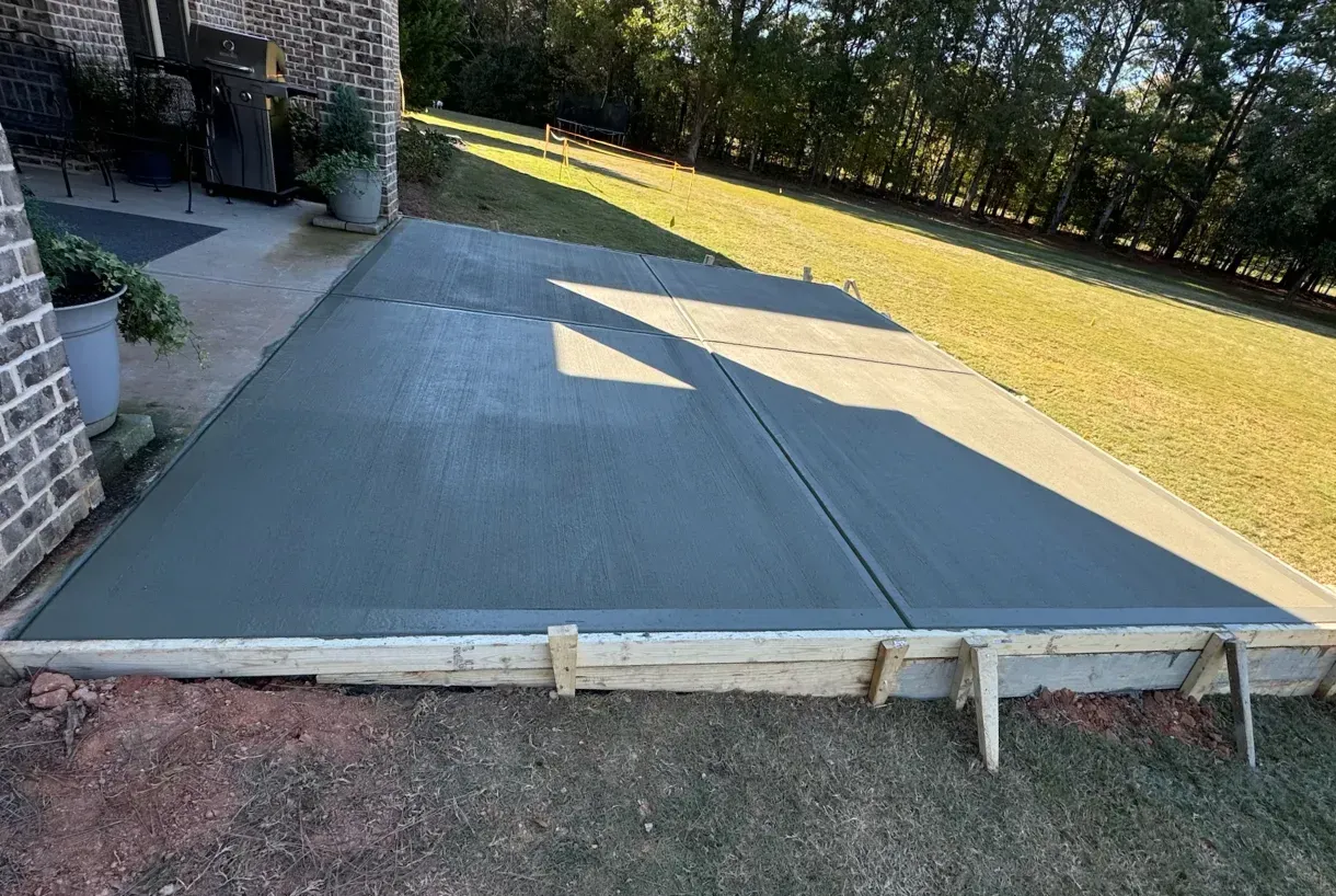 A newly poured gray concrete patio slab set in wooden forms next to a brick house in a backyard.