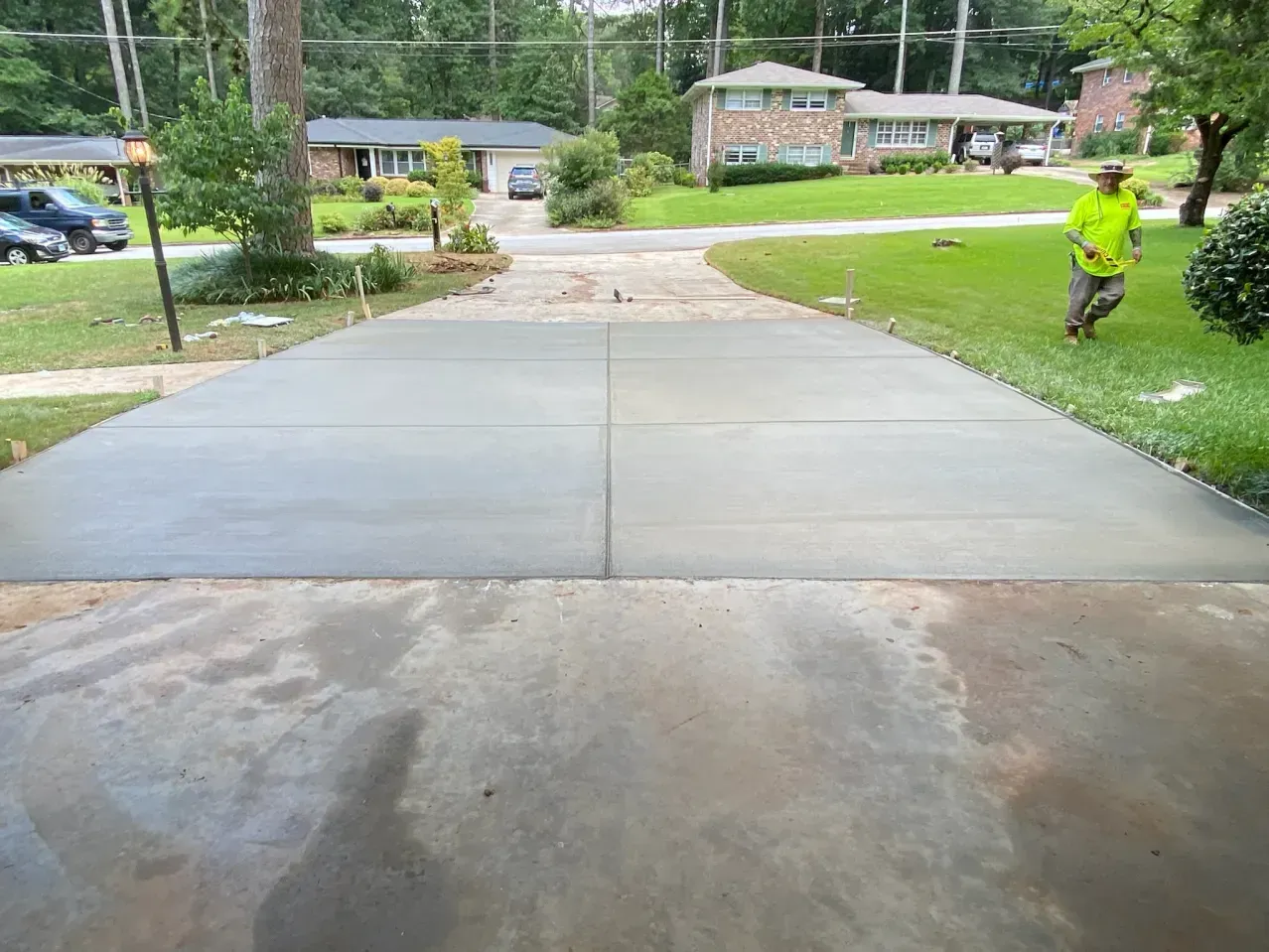 A freshly poured concrete driveway section in a residential front yard with a person in a bright yellow shirt standing by.