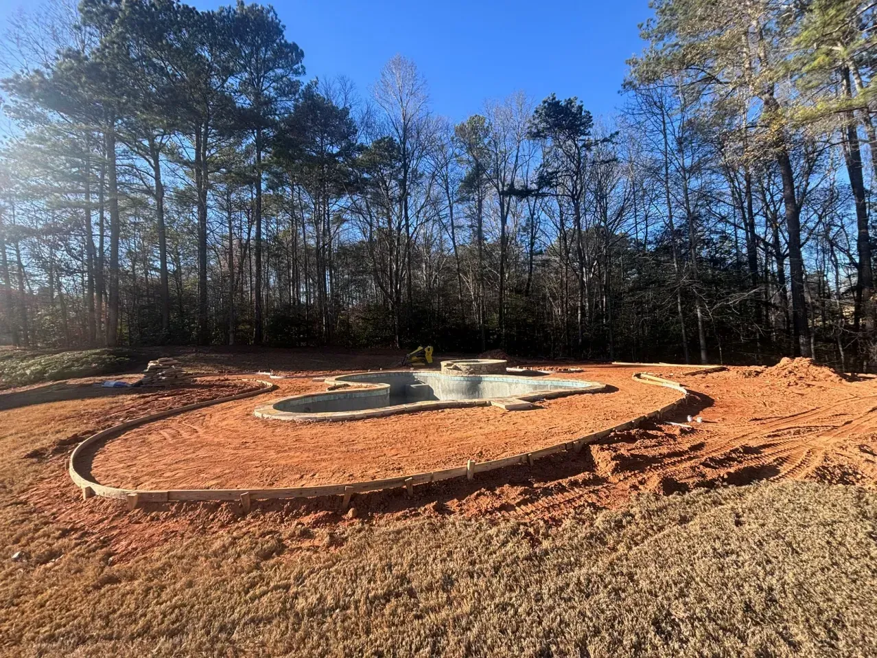 A concrete swimming pool shell under construction, surrounded by red dirt and a curved stone border in a wooded yard.