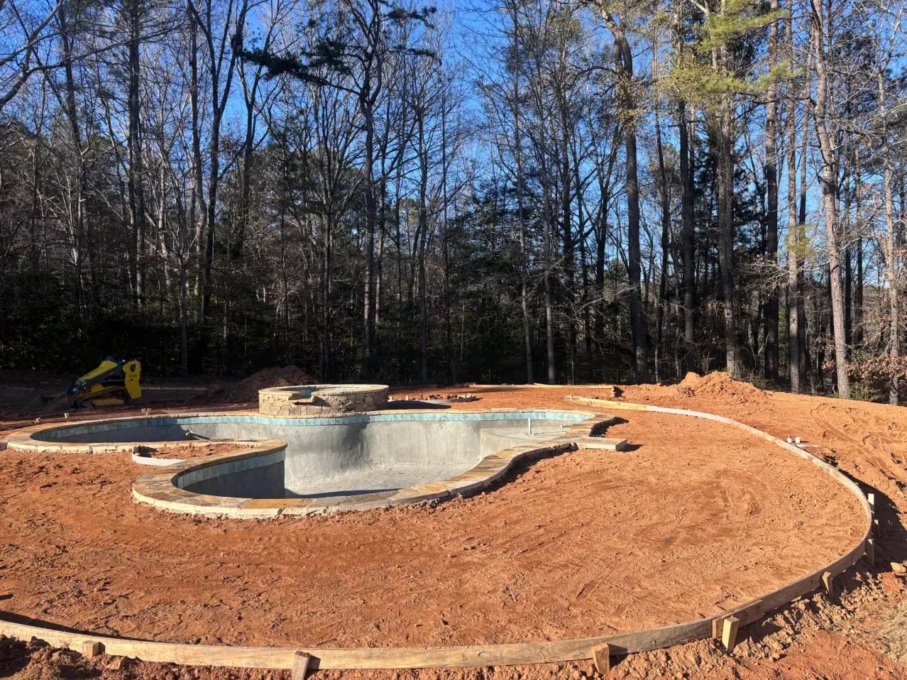 An in-ground swimming pool under construction, surrounded by bare red dirt and wooden framing for a deck.