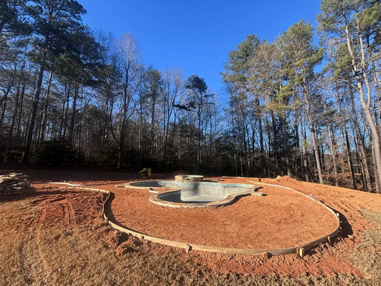 An empty, kidney-shaped pool under construction, surrounded by red dirt and wooden forms, set against a wooded backdrop.
