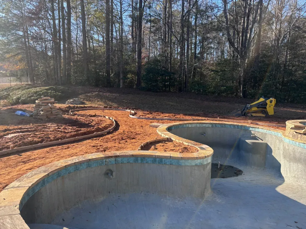 An empty, curved swimming pool sits in a yard under construction, surrounded by piles of red dirt and a skid steer loader.