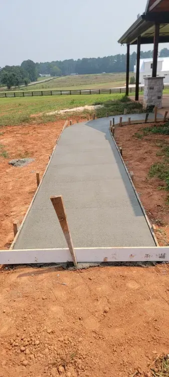 A freshly poured concrete sidewalk path leading toward a porch in a rural, grassy field setting.
