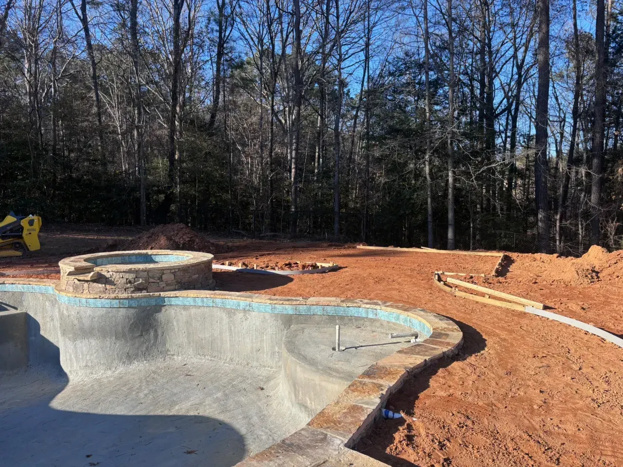 A half-finished swimming pool with a stone spillway and retaining wall under construction in a wooded backyard.