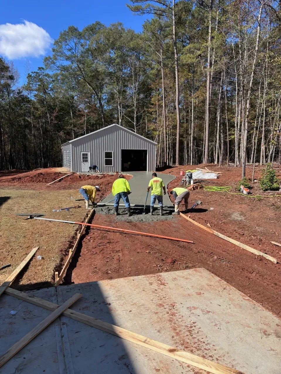 Workers in high-visibility vests smooth wet concrete for a new driveway in front of a metal garage building.