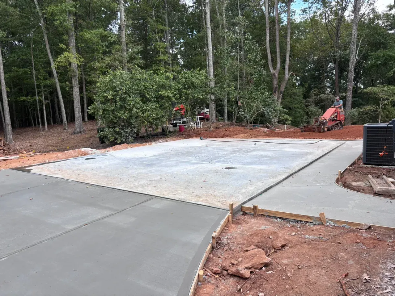 A newly poured concrete driveway and rectangular slab set against a background of trees and red soil.