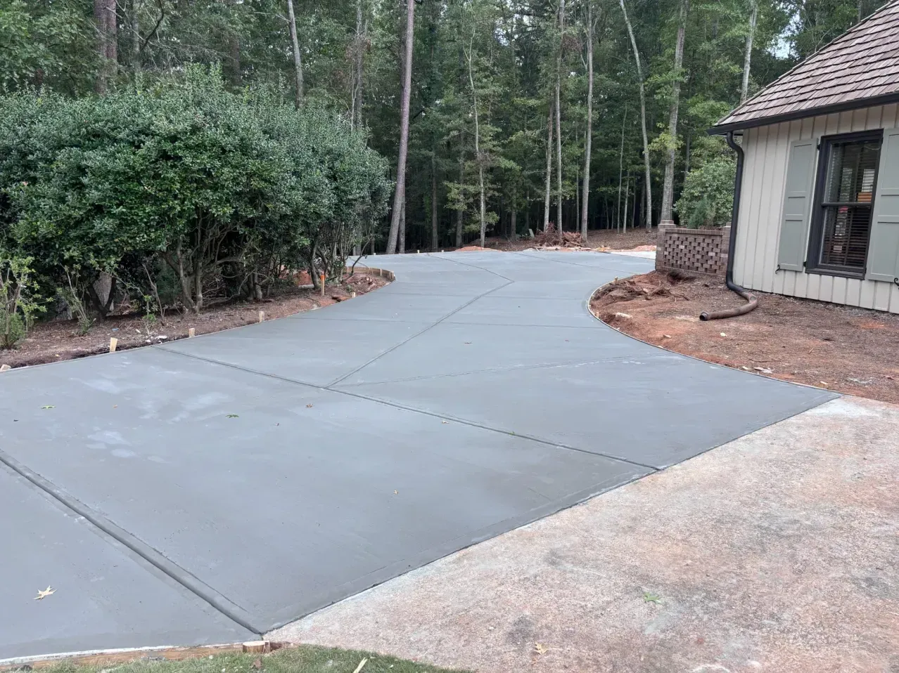 A freshly poured, smooth gray concrete driveway extending toward a wooded area next to a house with tan siding.