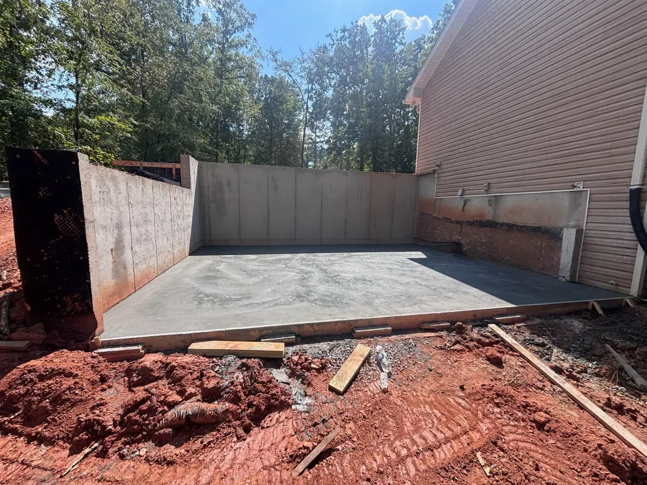A freshly poured concrete foundation and retaining walls next to a brick house under construction on a sunny day.