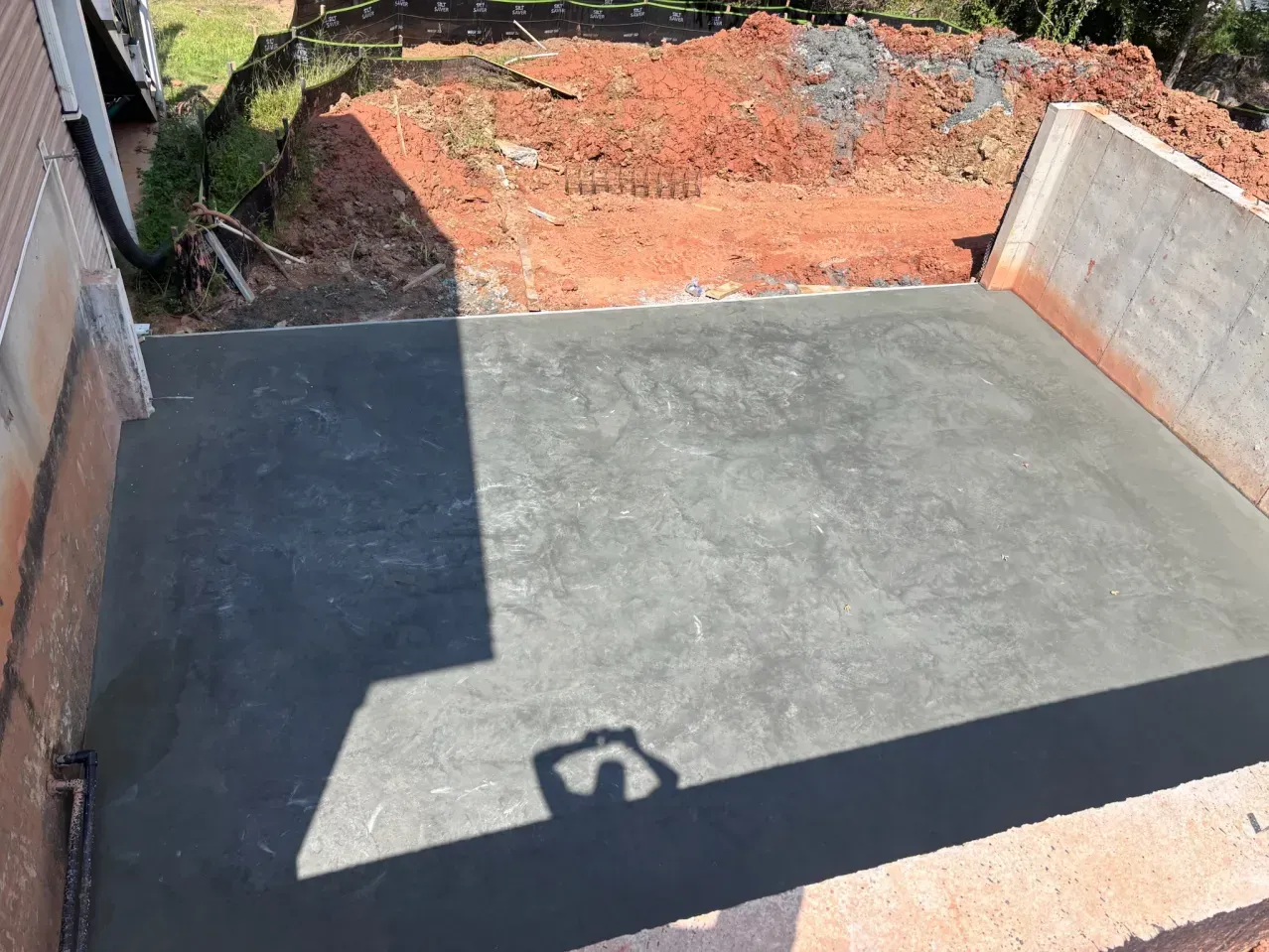A freshly poured concrete patio slab adjacent to a brick wall with a dirt excavation area in the background.