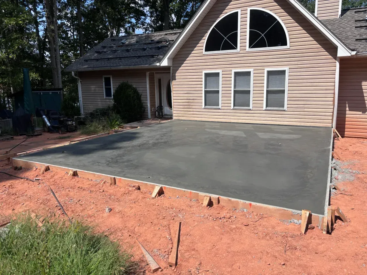 A freshly poured concrete patio slab in front of a house with light-colored siding and an arched window.