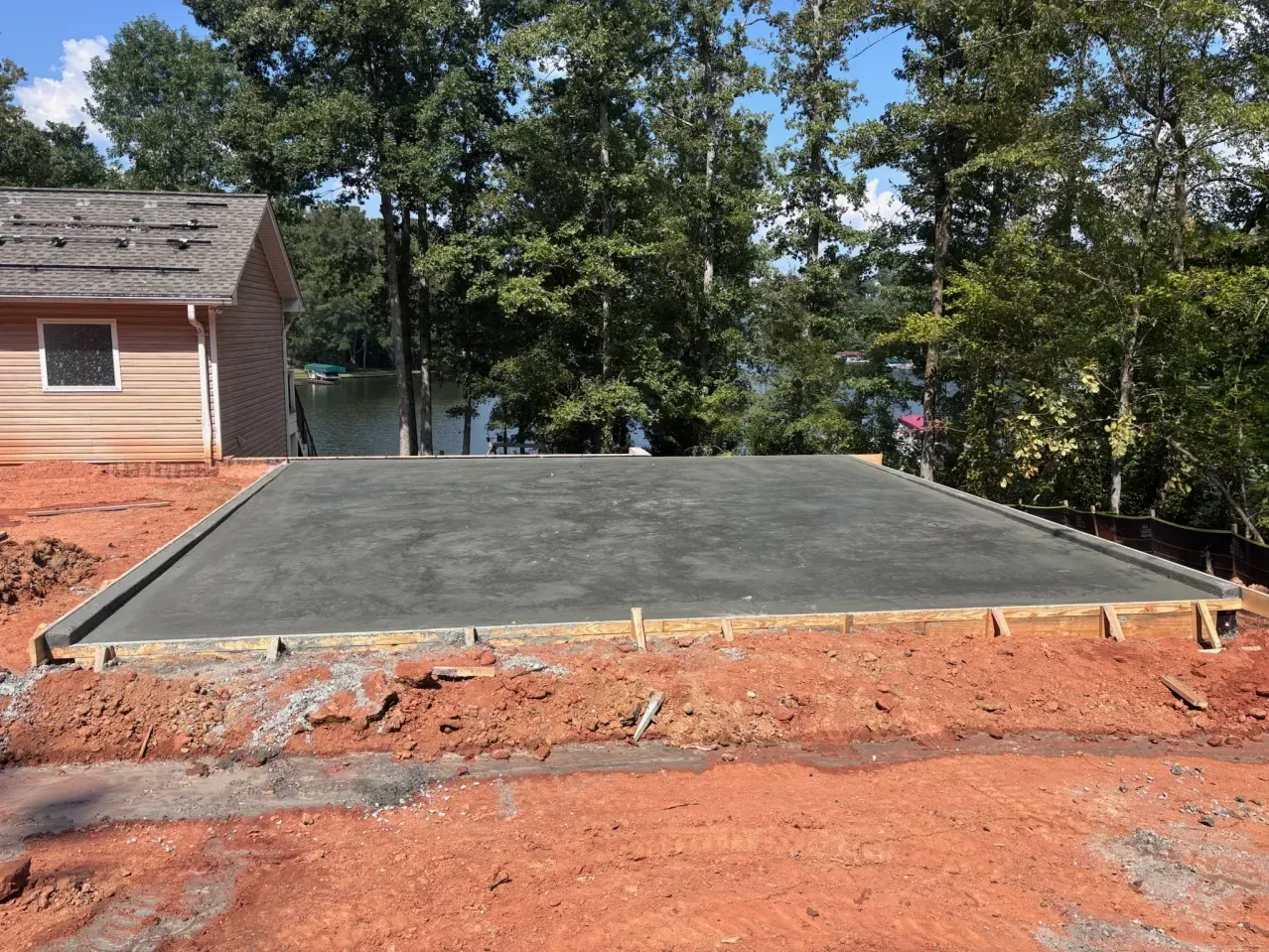 A newly poured concrete slab foundation sits in a yard near a house and trees, surrounded by red dirt.