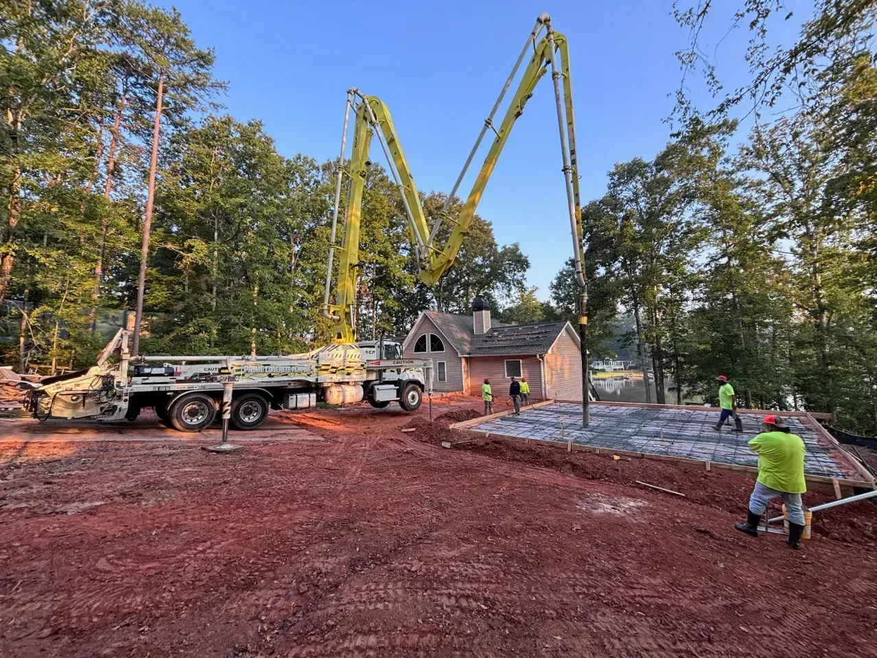 A bright green concrete pump truck pours wet concrete onto a foundation site near trees and a small house.