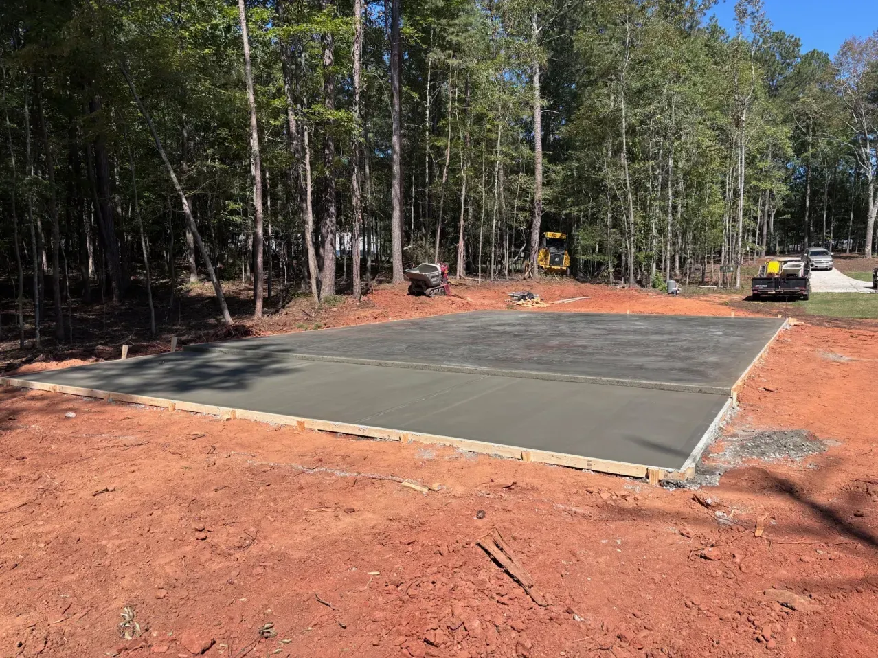 A freshly poured, rectangular concrete slab sits on red clay soil in a wooded clearing on a sunny day.