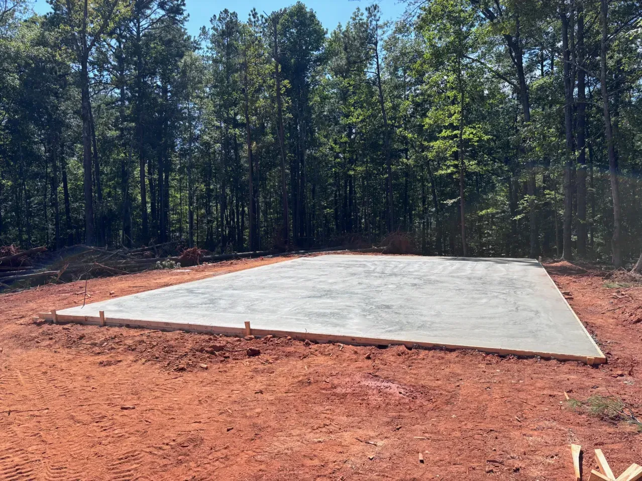 A freshly poured rectangular concrete slab foundation sits on red dirt surrounded by a lush forest under a clear sky.
