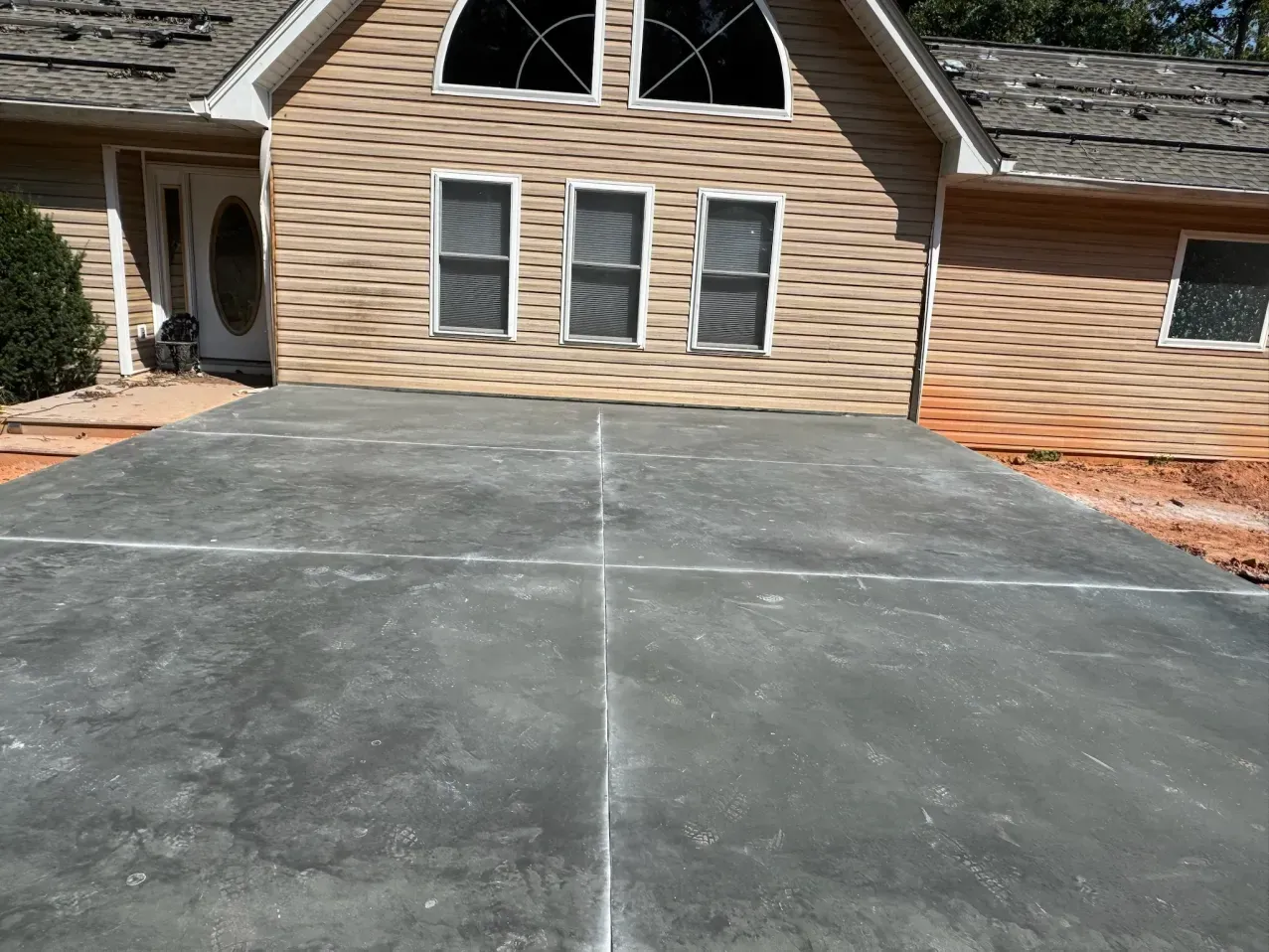 A new, gray concrete driveway with visible control joints sits in front of a tan-sided house with three windows.