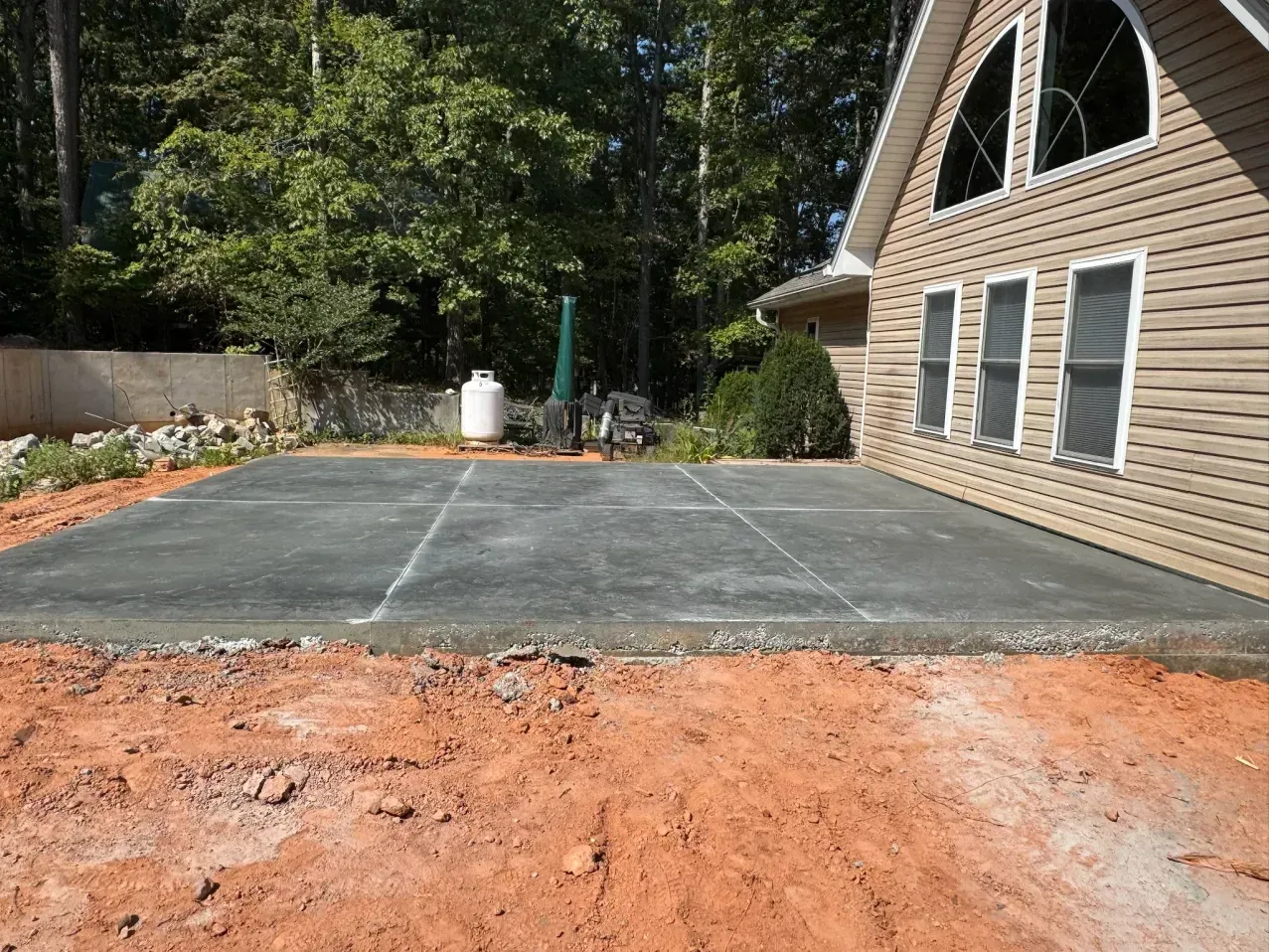 A newly poured, rectangular concrete slab sits next to the tan, vinyl-sided wall of a house with red clay soil in front.