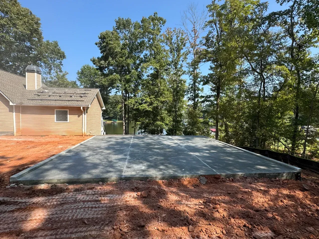 A newly poured rectangular concrete slab sits on red dirt beside a house near a wooded shoreline.