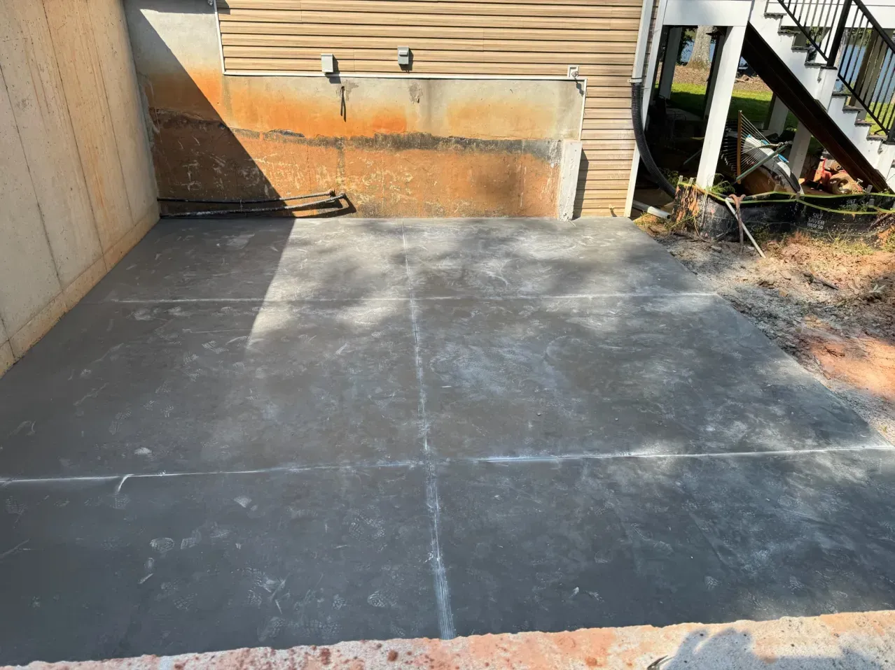 A newly poured gray concrete patio slab beside a house with beige siding and a wooden staircase.