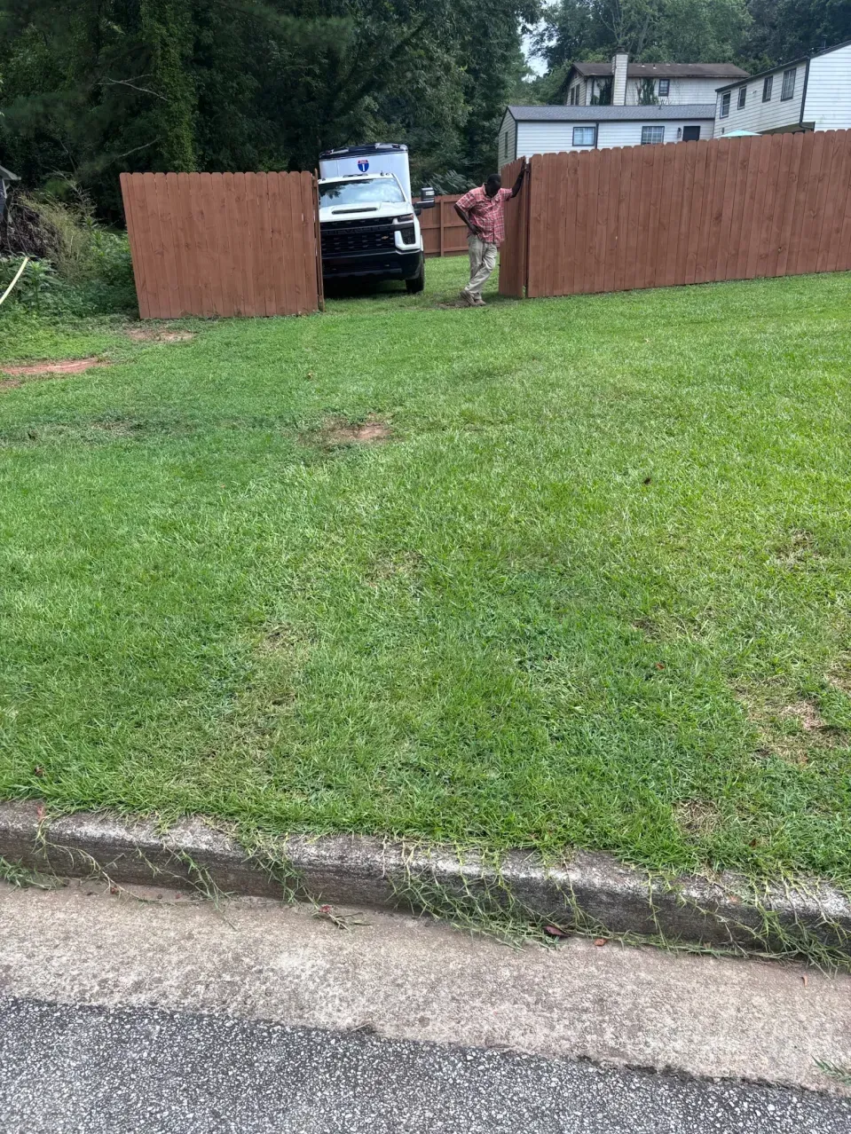 A person stands by a brown wooden fence in a grassy yard, with a white police vehicle parked behind the fence opening.
