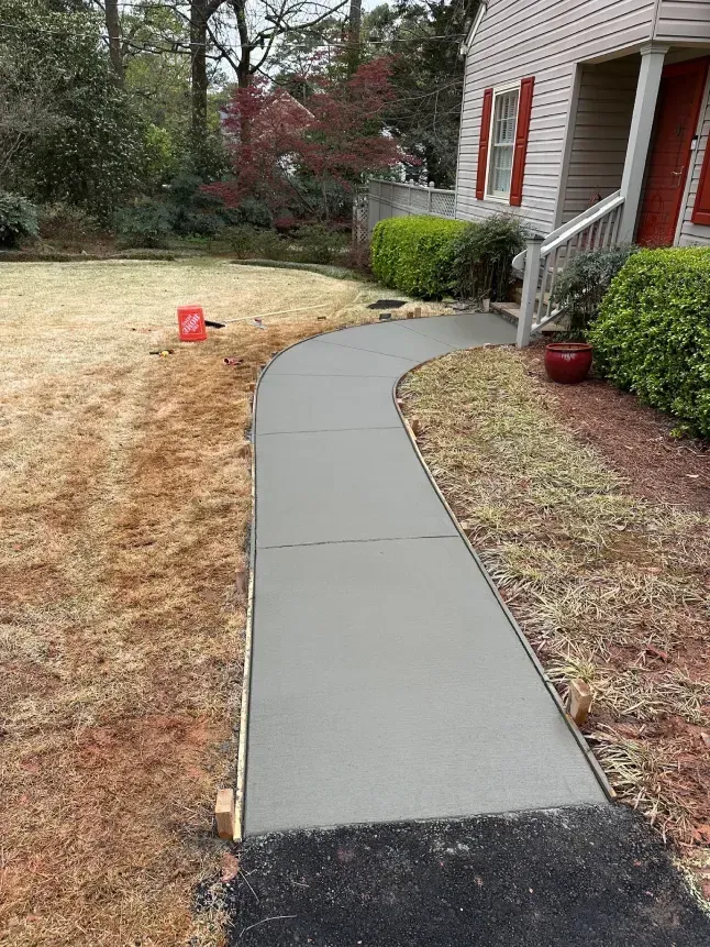 Curved concrete walkway beside a brick house, with shrubs and a dry lawn in front.
