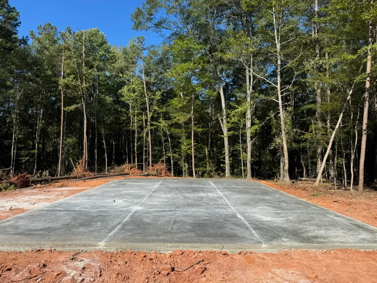 A rectangular concrete slab foundation situated on a red dirt clearing in front of a dense forest under a clear blue sky.