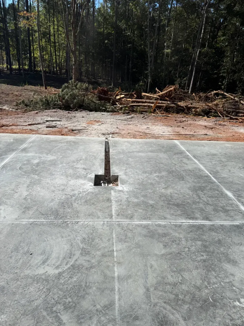 A metal post centered on a rectangular concrete slab, with a wooded background and cleared debris.