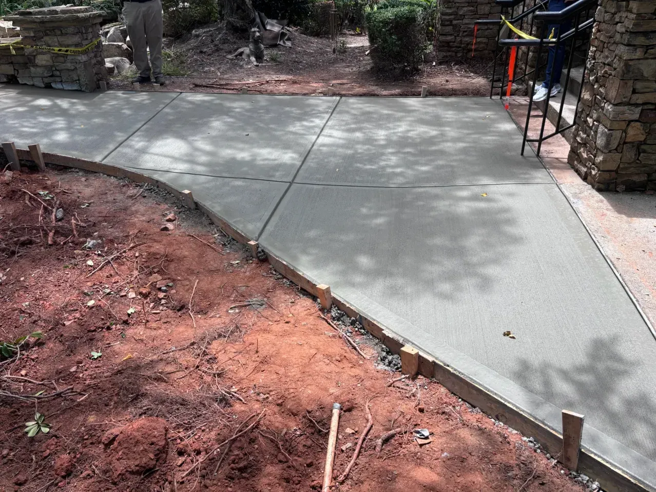 A newly poured concrete walkway curves past a stone pillar and earthen garden bed, with wooden form boards still in place.