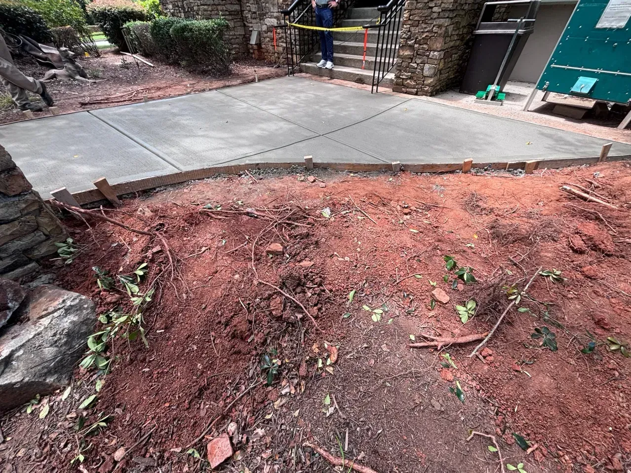 A newly poured concrete patio with wooden border forms, located in front of a stone building with red soil in the foreground.