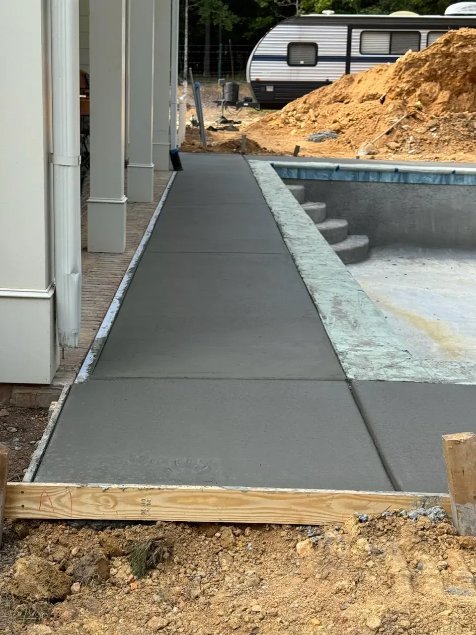 Newly poured gray concrete deck alongside a swimming pool under construction, with a trailer and dirt pile in the background.