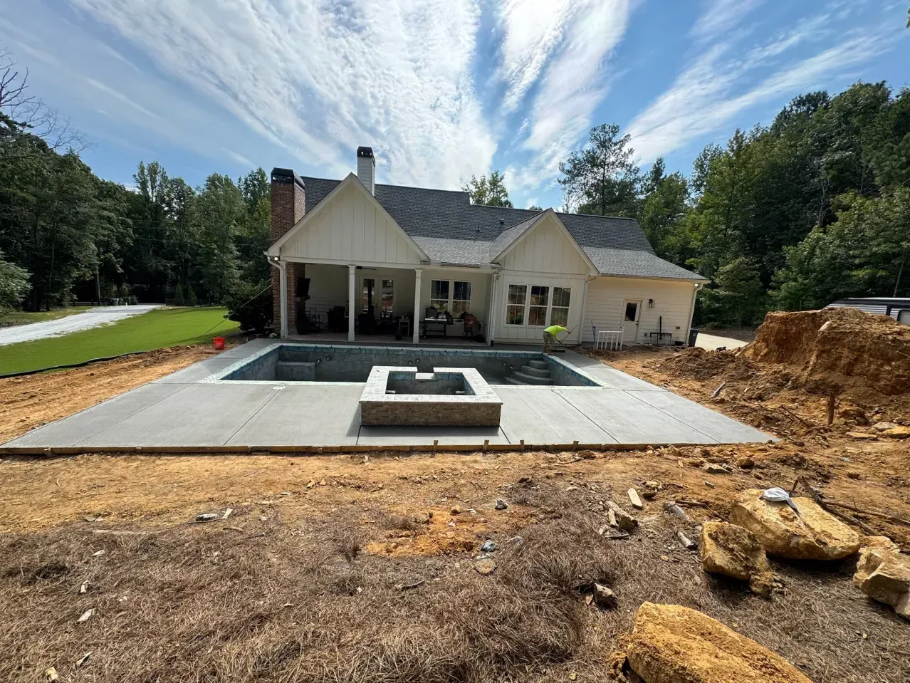 A backyard pool and stone fire pit under construction behind a white house with a partially finished concrete patio.