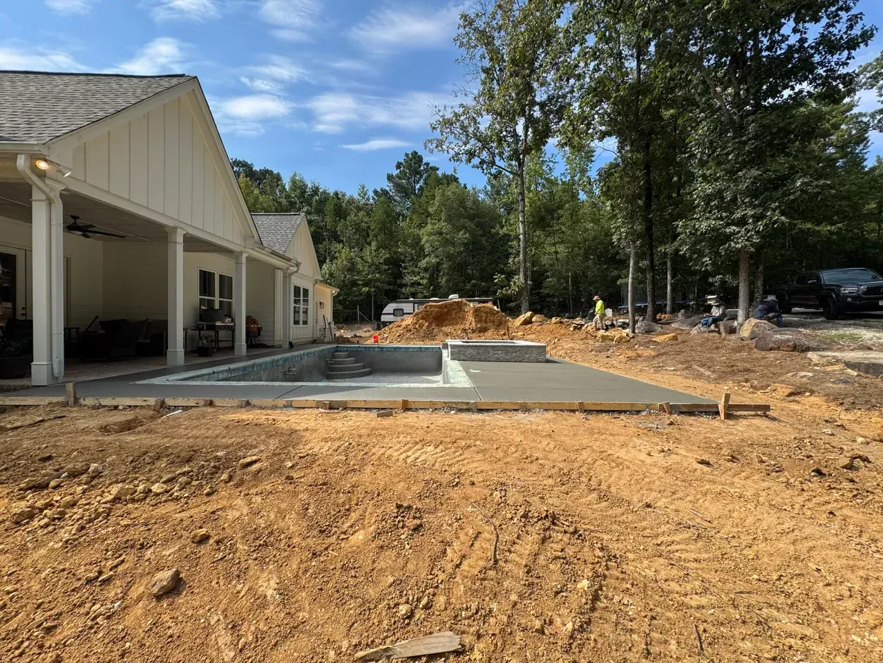 A new concrete pool deck and pool shell are being installed next to a house with a covered porch on a dirt construction site.