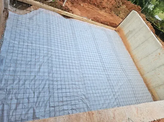 A construction site showing a concrete foundation base covered with plastic sheeting and a steel wire mesh grid.