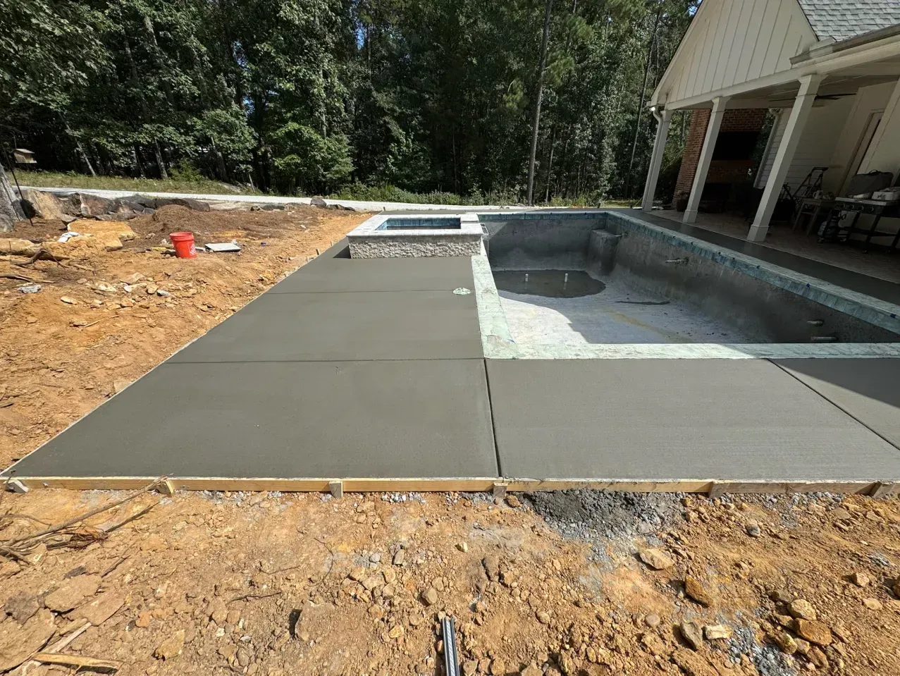 Freshly poured concrete patio surrounding an empty swimming pool and spa under construction on a sunny day.