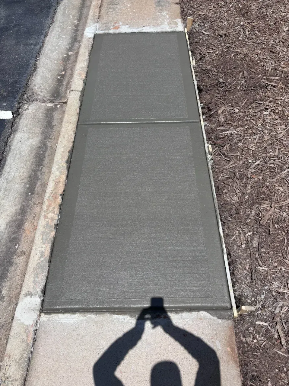 A newly poured rectangular concrete sidewalk section between an existing path, a paved curb, and a mulch bed.