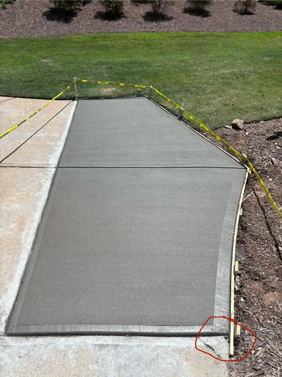 A newly poured concrete walkway section under construction, marked by yellow caution tape and a wooden form board.