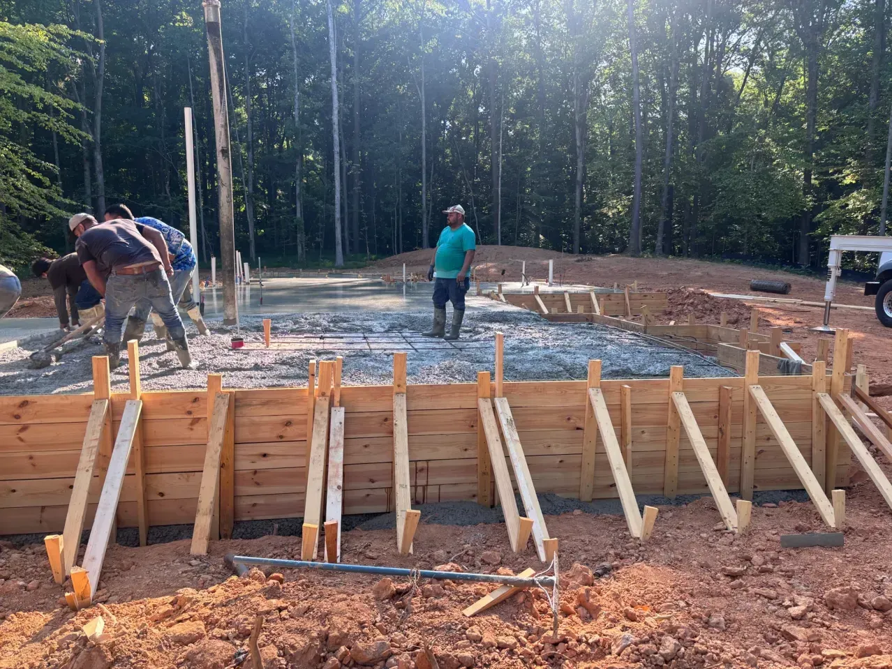 Construction workers prepare a gravel building foundation surrounded by wooden formwork in a forest clearing.