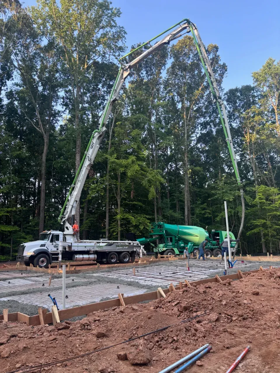 A concrete pump truck with a long boom arm extends over a construction site with a mixer truck and gravel foundation.