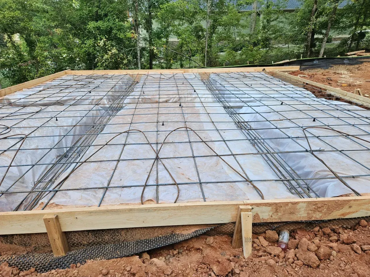 Wooden formwork contains a rebar grid atop a plastic vapor barrier for a concrete foundation on an outdoor dirt site.