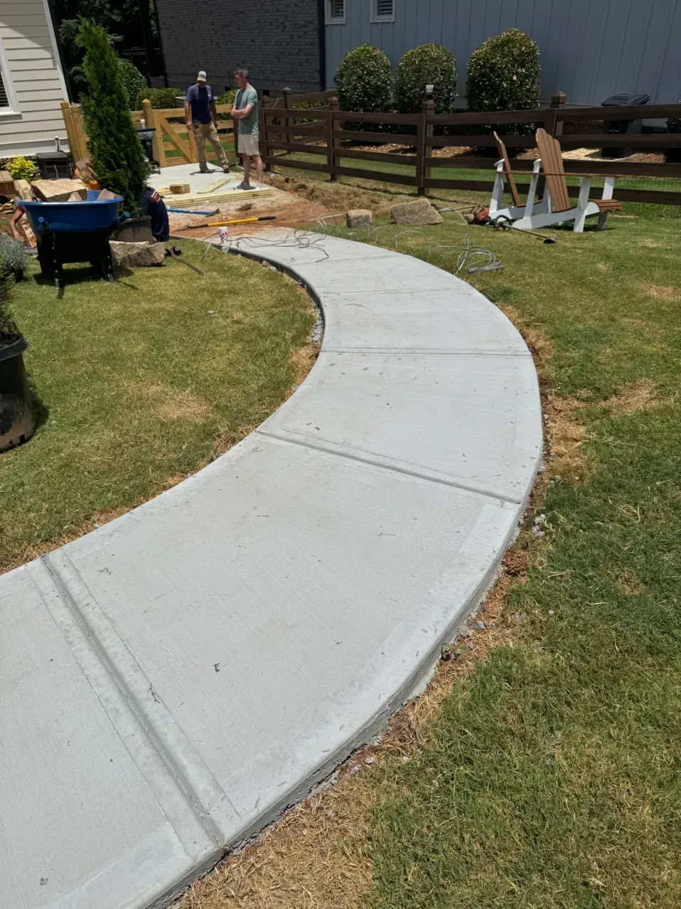 A newly poured concrete walkway curves through a grassy yard toward two people working in the background.