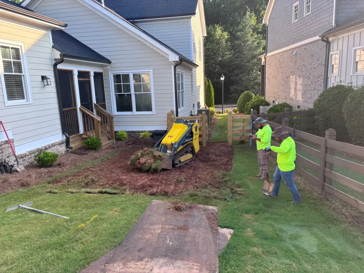 Two workers in neon shirts operate a skid steer to landscape a yard next to a suburban house.