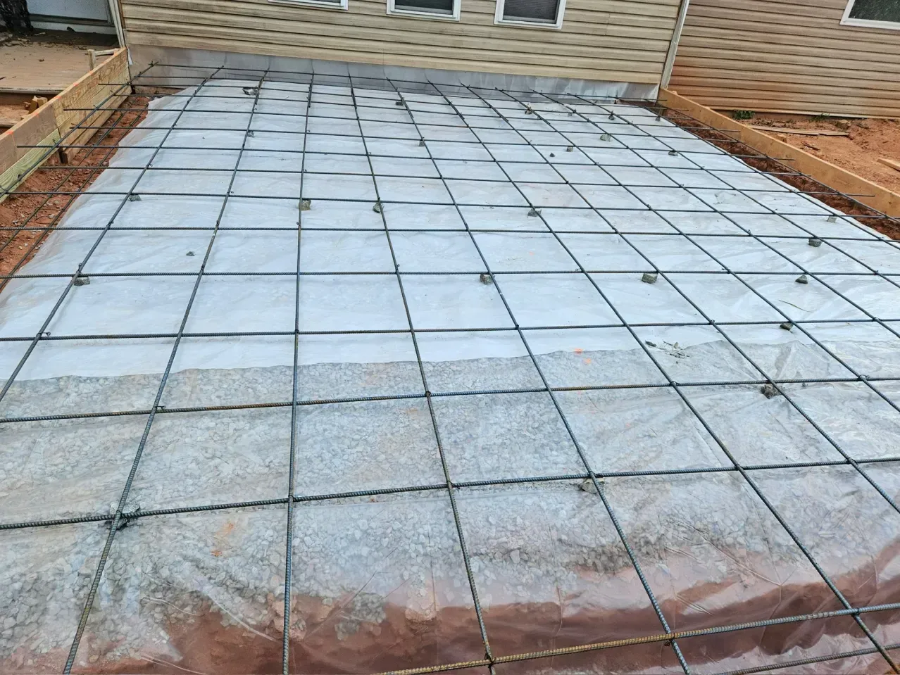 A concrete patio project site with a wire mesh grid laid over a vapor barrier between wooden forms next to a house.